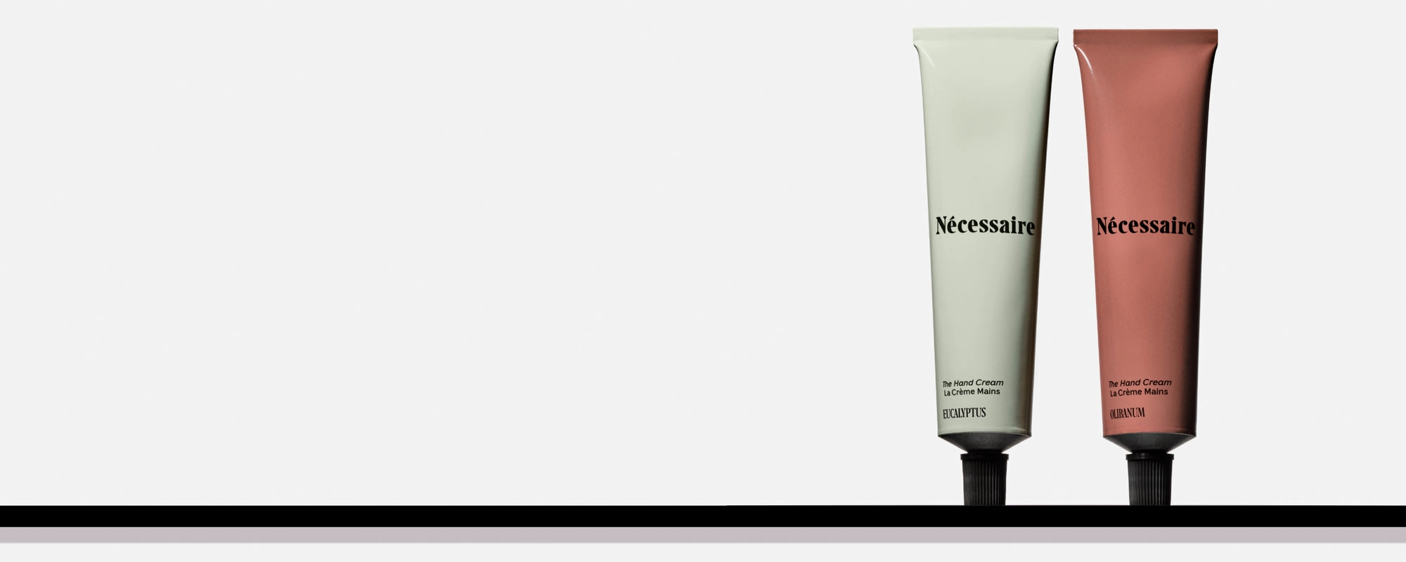 Two Nécessaire hand cream tubes, one pale green and one terracotta, standing on a black shelf against a soft grey wall